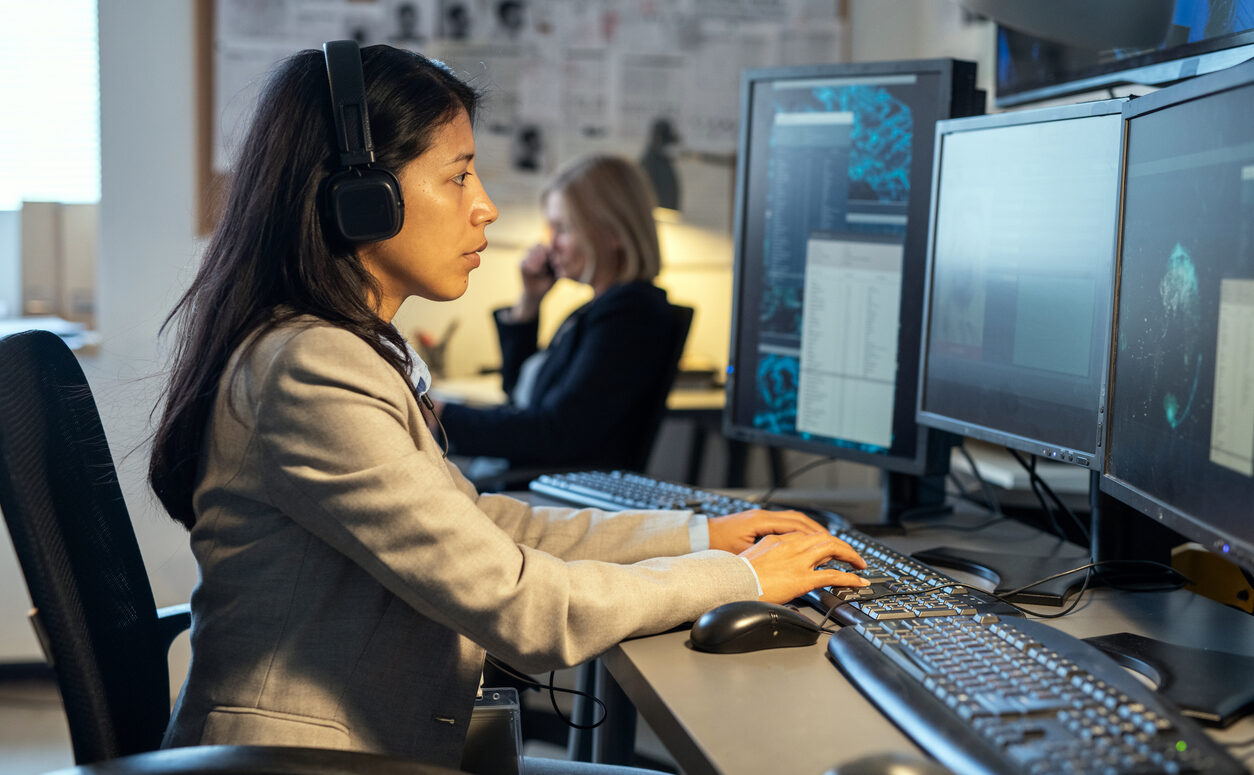 Side view of young Hispanic female operator of surveillance system Side view of young Hispanic female operator of surveillance system sitting by desk in front of computer monitors in office