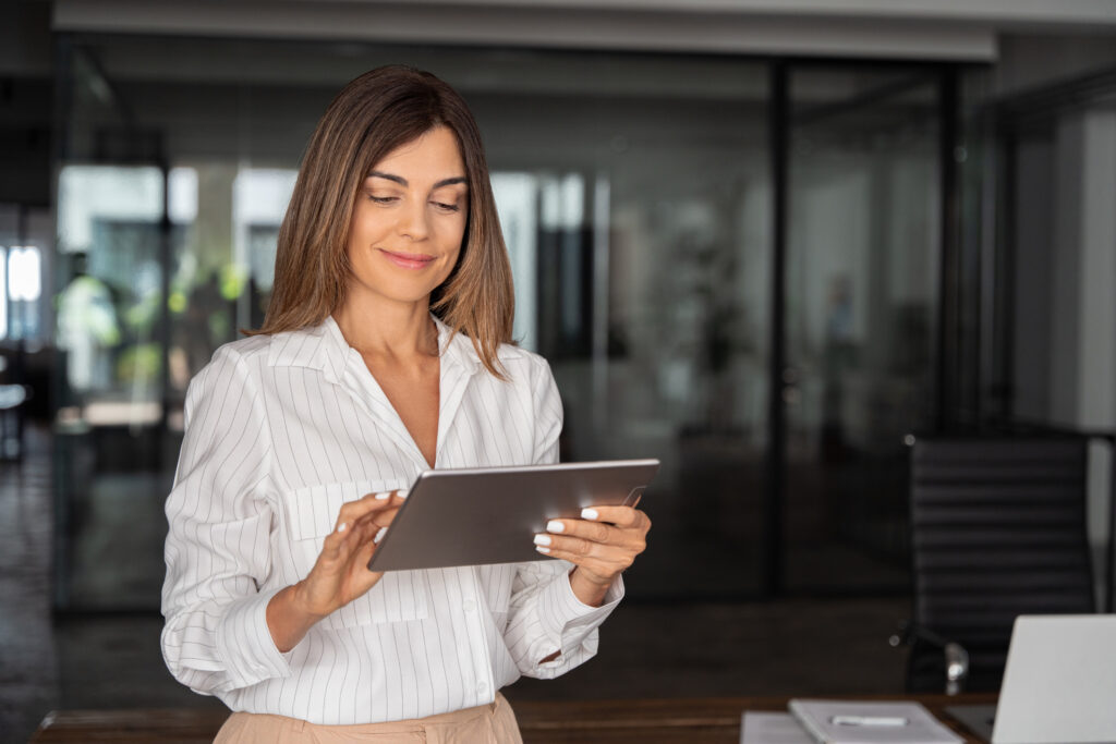 Latin Hispanic mature adult professional business woman looking at online trade app. European businesswoman CEO holding digital tablet using fintech tab application standing at workplace in office.